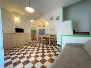 A compact living space with a checkered floor, a dining area with a table and chairs, a kitchenette with cabinets and a TV, and a green-framed window. - Bright Two-storey Flat (San Michele al Tagliamento)