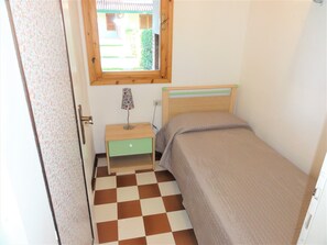 Een kleine kamer met een enkel bed, een geruit vloertje, een nachtkastje met lamp en een raam met uitzicht op een tuin.
