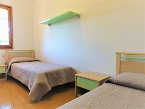 Quarto simples com duas camas, uma estante verde e uma janela.