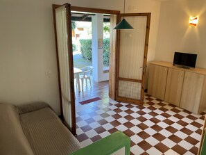 A room with a checkered floor, a green bench, a TV on a wooden cabinet, and a door leading to an outdoor area with a table and chairs. - Bright Two-storey Flat (San Michele al Tagliamento)