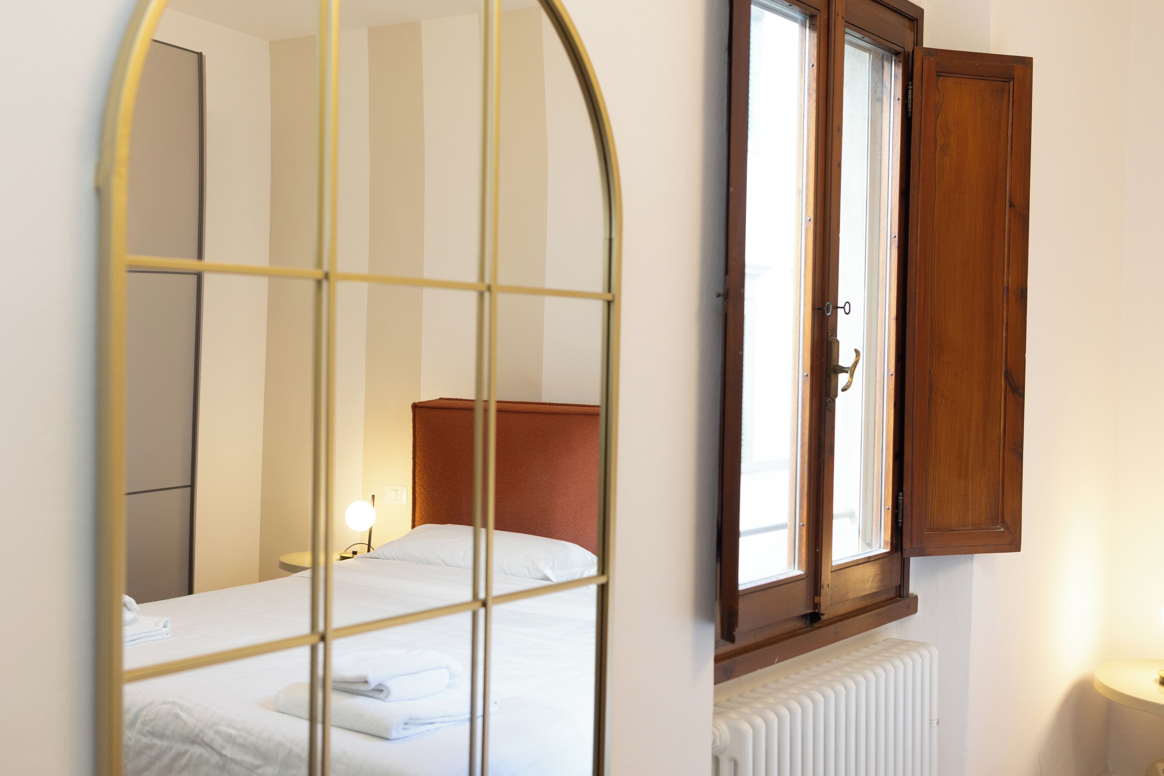 A bedroom with a bed, a radiator, a window with a wooden shutter, and a mirror with a grid pattern.