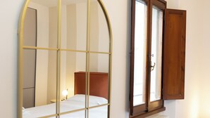 Une chambre à coucher comprenant un lit, un radiateur, une fenêtre avec un volet en bois et un miroir à motifs géométriques.