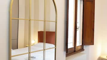 A bedroom with a bed, a radiator, a window with a wooden shutter, and a mirror with a grid pattern.