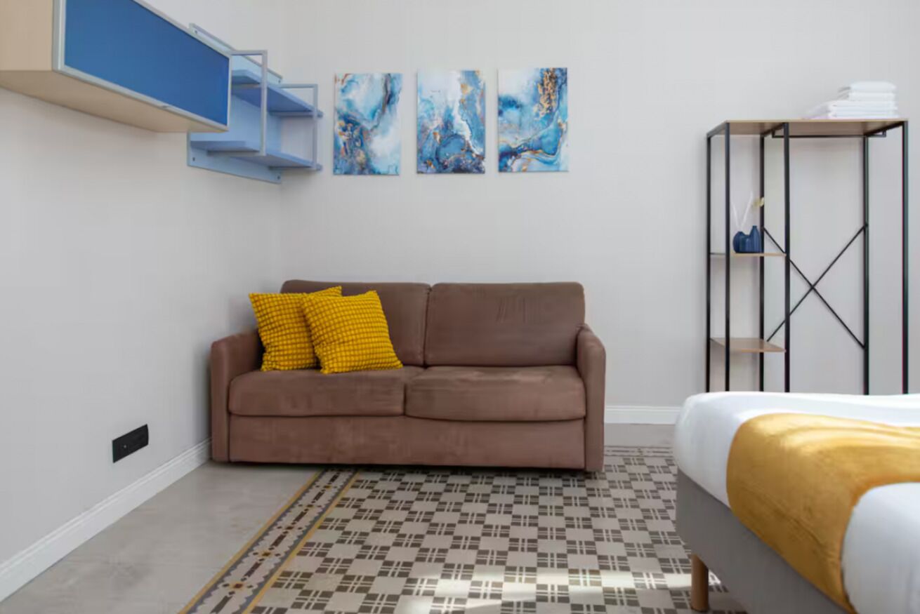 Une chambre moderne avec un canapé marron, un tapis à motifs, un lit agrémenté d’un couvre-lit doré et des œuvres d’art abstraites accrochées au mur.