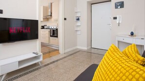 Ein modernes Wohnzimmer mit Flachbildfernseher, einer gelben Kissen ausgelegten Couch und einer Küche im Hintergrund, sowie einer weißen Tür.