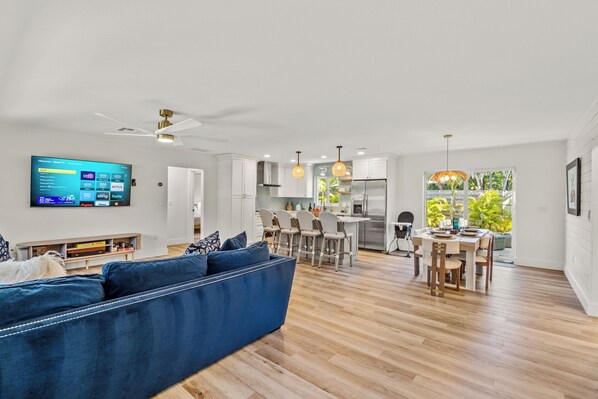 A spacious living area with a blue sofa, wooden flooring, a dining table with chairs, and a kitchen in the background.
