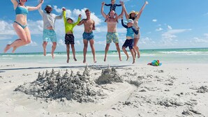 Une plage de sable où l’on voit des gens sauter, et un château de sable au premier plan.