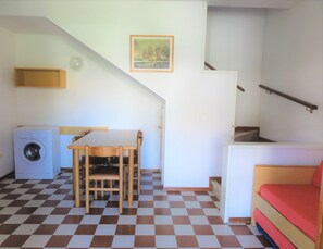 Una stanza compatta con pavimento a scacchi, una lavatrice, un tavolino con sedie, un letto e una scala.