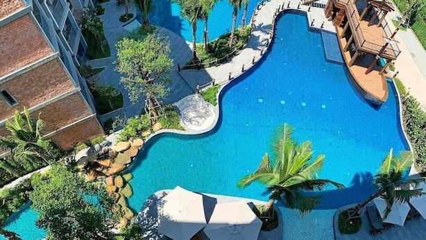 A large, winding pool with palm trees and lounge chairs, surrounded by a building with balconies. - Pool Side Retreat- 1 BED TC-B105 (Sa Khu)