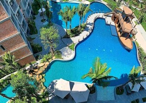 A large, winding pool with palm trees and lounge chairs, surrounded by a building with balconies. - Pool Side Retreat- 1 BED TC-B105 (Sa Khu)