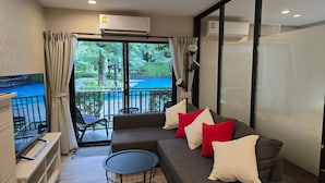 A modern living room with a grey sofa, red and white cushions, a small round table, and a view of a pool and greenery outside. - Pool Side Retreat- 1 BED TC-B105 (Sa Khu)