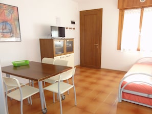 Une pièce avec un bureau en bois, des chaises blanches, un lit avec un matelas rose, une porte en bois et une fenêtre ornée de rideaux en dentelle.