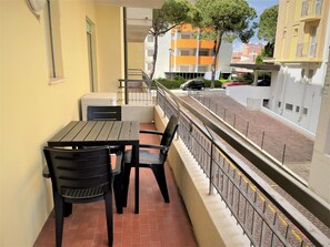 A balcony with a table and chairs, a railing, and a view of a street with parked cars and buildings. - Cozy Apartment Close to the Beach (San Michele al Tagliamento)