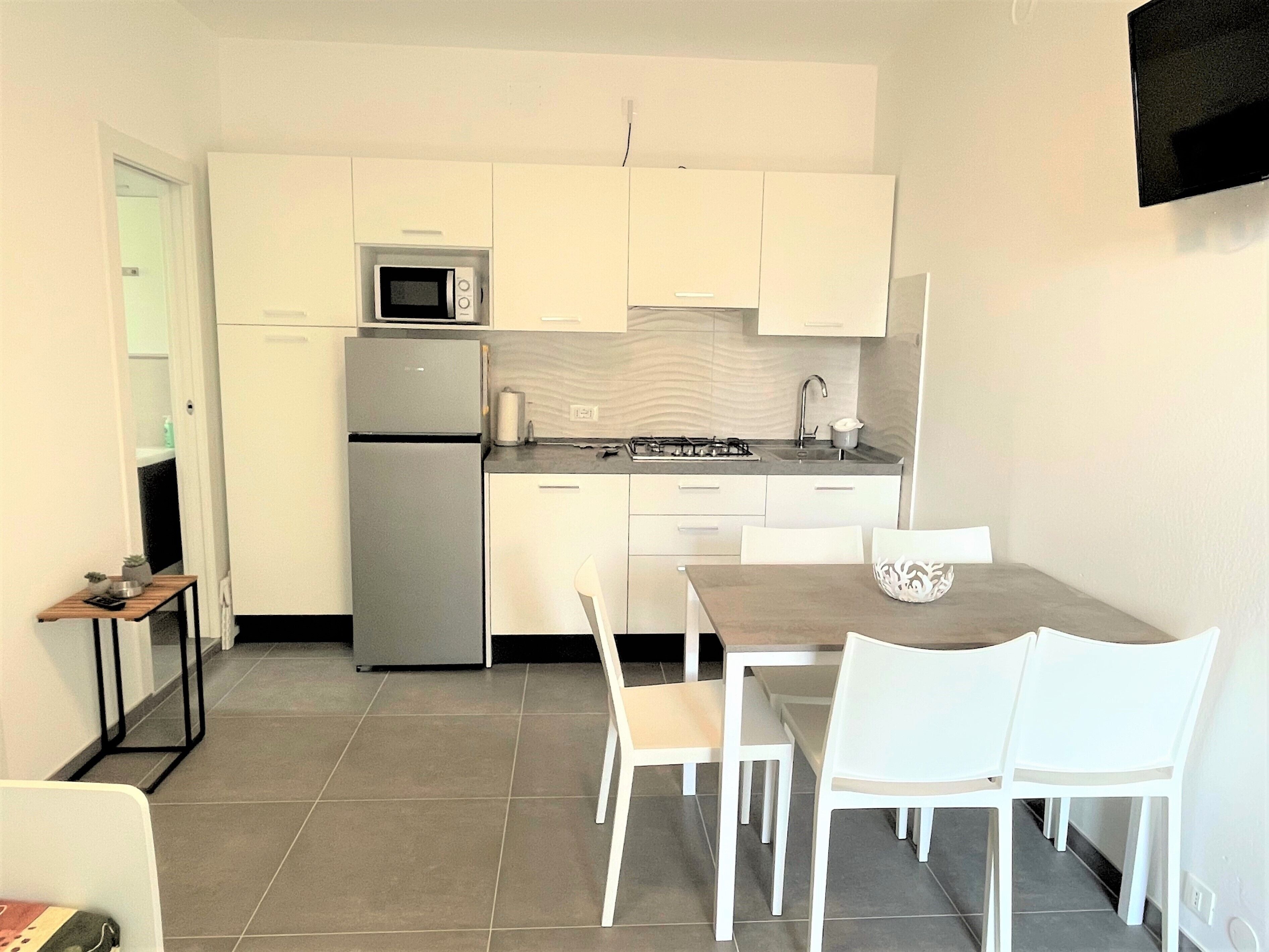 A compact kitchen with white cabinets, a microwave, a refrigerator, a sink, and a dining area with white chairs.