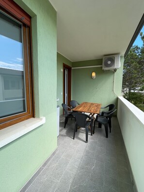 Un balcón con mesa de madera y cuatro sillas, una ventana con persianas y un equipo de aire acondicionado.