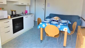 A compact kitchen with white cabinets, a stove, and a sink. There is a dining table with a blue tablecloth and four chairs. A blue sofa is against the wall. - Holiday Home Near the Sea-beahost (San Michele al Tagliamento)