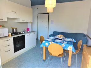 A compact kitchen with white cabinets, a stove, and a sink. There is a dining table with a blue tablecloth and four chairs. A blue sofa is against the wall.