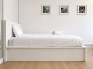 Een minimalistisch wit bed met een kussen en een opgerolde handdoek, voor een effen muur met drie ingelijste kunstwerken.