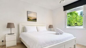 Een moderne slaapkamer met een wit bed, twee nachtkastjes, een wandlamp en een schilderij aan de muur, met een raam dat uitkijkt op groen.