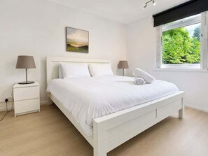 Een moderne slaapkamer met een wit bed, twee nachtkastjes, een wandlamp en een schilderij aan de muur, met een raam dat uitkijkt op groen.