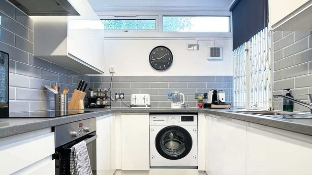 Een moderne keuken met witte kastjes, een ingebouwde wasmachine, een klok aan de muur en een raam boven de gootsteen.