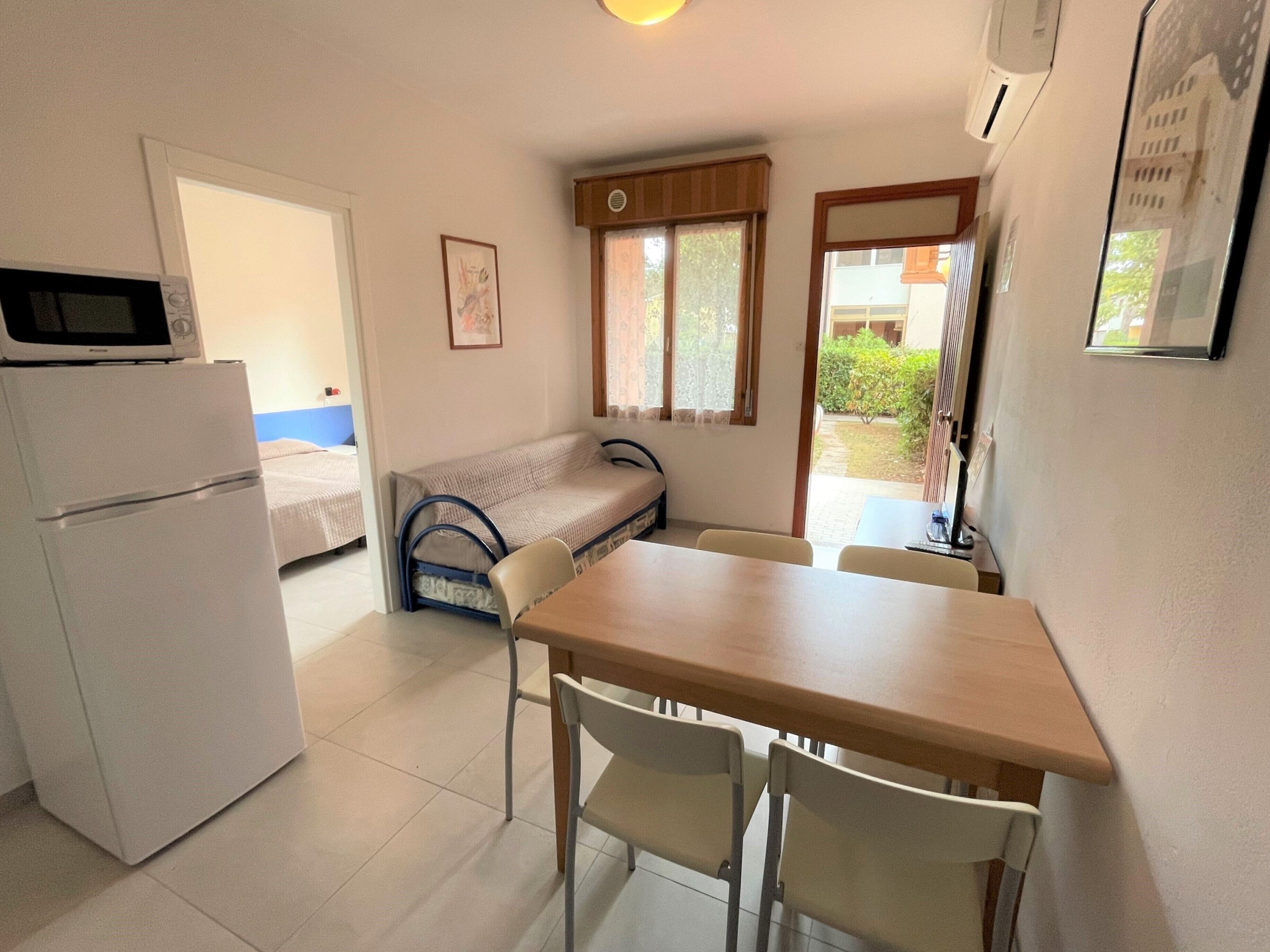 Une pièce compacte comprenant un coin cuisine, un lit simple, une petite table avec deux chaises et une fenêtre avec des rideaux.