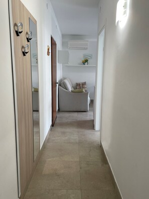 Ein Flur mit einer Holztür, einer weißen Wand, einem weißen Sofa mit Kissen und einem kleinen Regal mit Blumen.