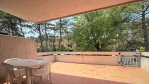 A covered outdoor patio with a table and chairs, surrounded by a low wall and trees. - Apartment With Private Entrance in Bibione (San Michele al Tagliamento)