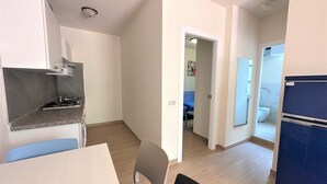 A compact kitchen with a stove, a dining table with chairs, and a refrigerator. There is a hallway leading to a bedroom with a bed and a bathroom with a toilet. - Apartment With Private Entrance in Bibione (San Michele al Tagliamento)