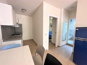 A compact kitchen with a stove, a dining table, and a chair. There is a hallway leading to a bathroom with a toilet and a shower. - Apartment With Private Entrance in Bibione (San Michele al Tagliamento)