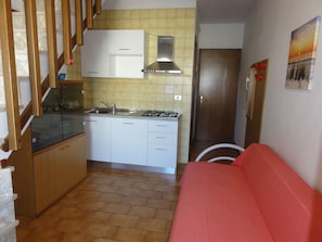 Cozinha compacta com fogão a gás, pia e armários brancos. Uma sala adjacente possui um sofá vermelho.