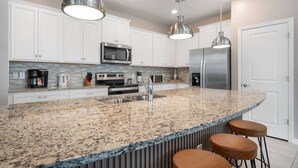 Una cocina moderna con armarios blancos, electrodomésticos de acero inoxidable, encimera de granito e iluminación colgante.