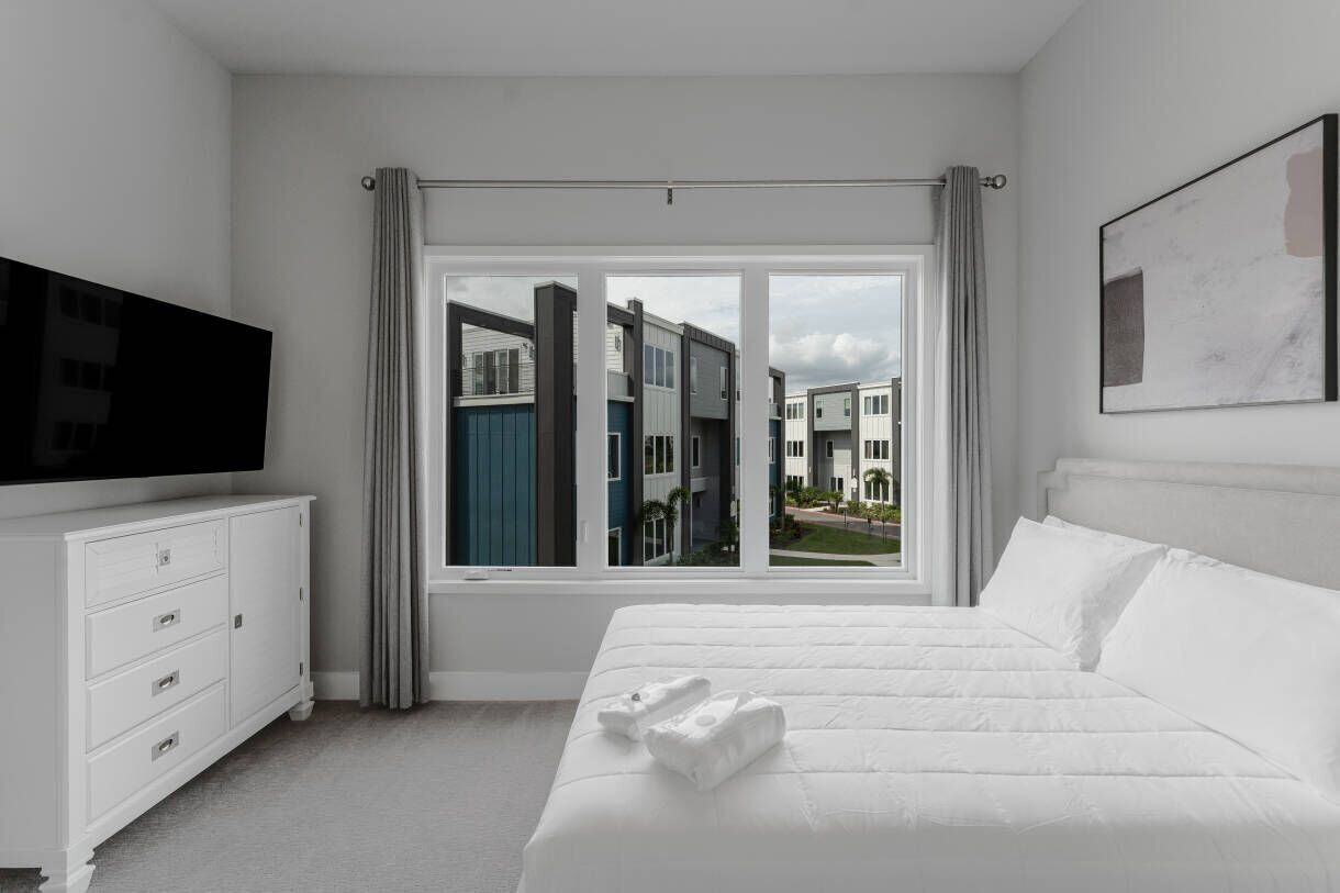 A hotel room with a large bed, a TV on a white dresser, and a view of neighboring buildings through a window.