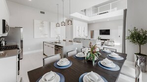A modern kitchen with a built-in oven, a dining area with a dark wooden table set for four, and a living area with a sofa and a TV. - Modern 2-Story Loft w/ Pool Table – Near Disney (Kissimmee)