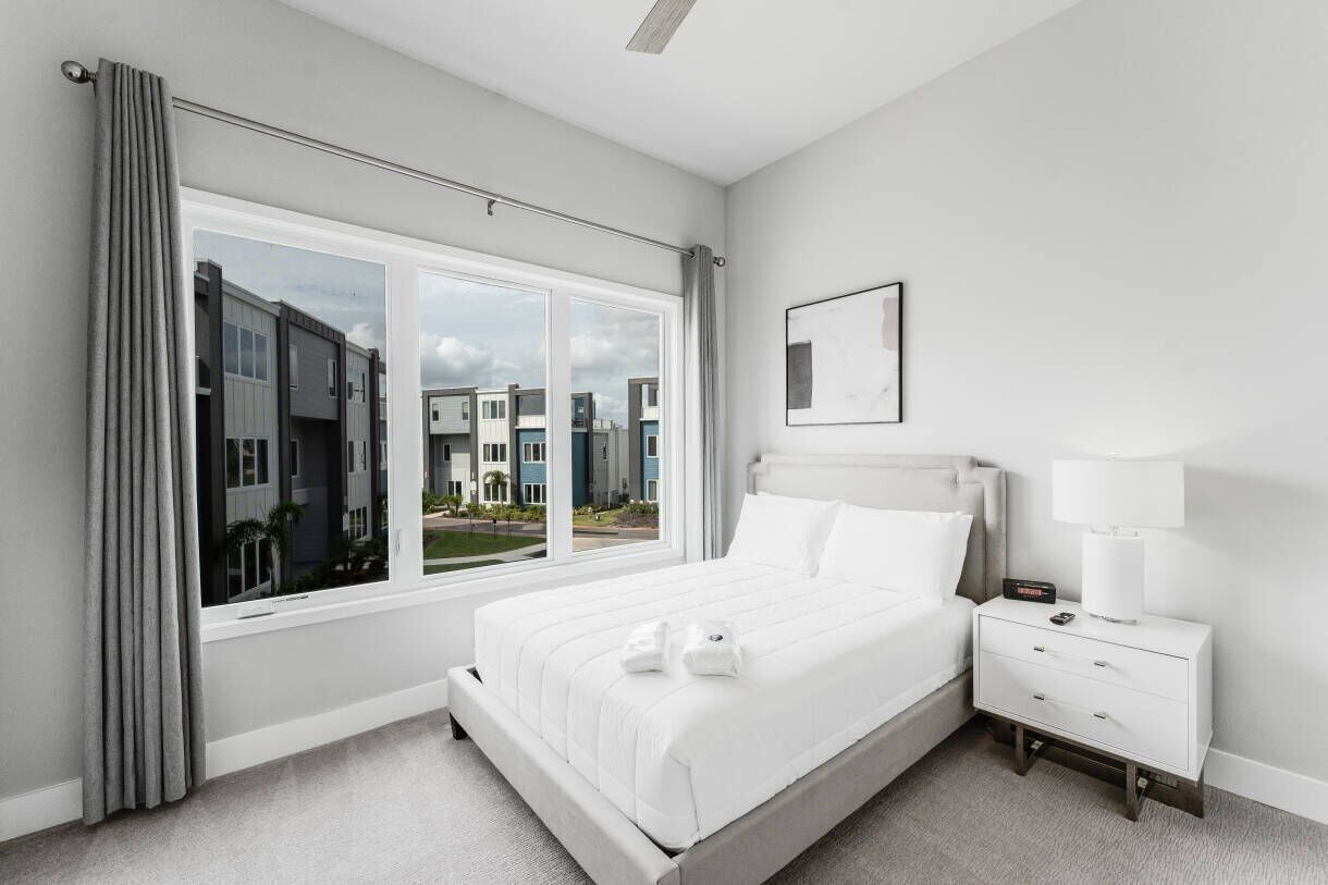 A modern hotel room with a large window, a neatly made bed, a bedside table with a lamp, and a framed picture on the wall.