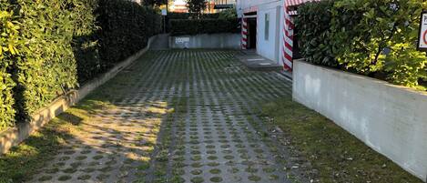 Un sendero pavimentado con diseños circulares, bordeado por setos y un edificio blanco con barreras a rayas rojas y blancas.