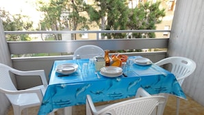 Une table à manger dressée pour quatre personnes sur un balcon, avec une nappe bleue, des chaises blanches et une vue sur les arbres et les bâtiments en arrière-plan.