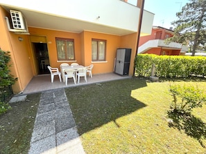 A residential building with a patio area featuring a table and chairs, a door, and a small garden with a hedge. - Cozy Flat in a Villa With Shared Pool (San Michele al Tagliamento)