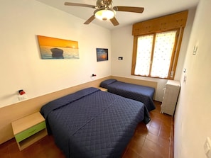 A bedroom with two beds, a ceiling fan, a window with curtains, and a painting on the wall. - Cozy Flat in a Villa With Shared Pool (San Michele al Tagliamento)