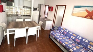 Een kleine keuken met een eettafel en stoelen, een kitchenette met een magnetron, en een bank met een decoratief kussen.