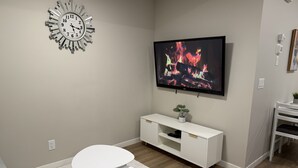 A modern living room with a flat-screen TV, a decorative clock, a white console table, and a small potted plant. - Cozy Basement Suite in Mahogany (Calgary)