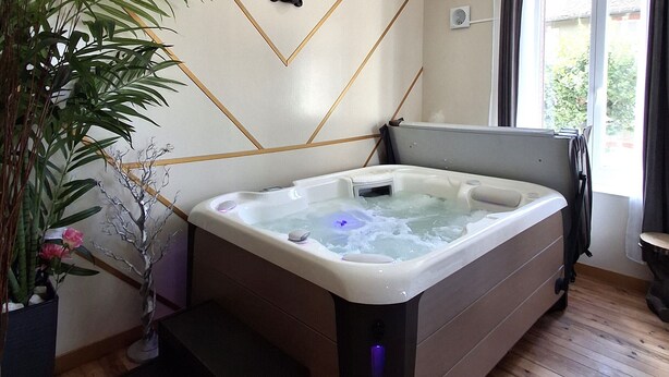 A modern bathroom with a freestanding hot tub, wooden flooring, a wall-mounted air conditioning unit, and a large window with curtains. - Le Bali Zen, studio with jacuzzi (Charavines)