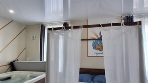 A bedroom with a bed, a small sofa, a hot tub, and a window with curtains. - Le Bali Zen, studio with jacuzzi (Charavines)