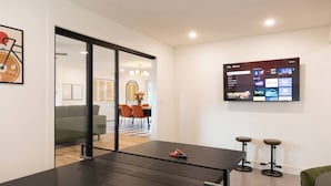 Une pièce moderne équipée d’une table de ping-pong, d’un téléviseur diffusant des services de streaming et d’un coin repas à l’arrière-plan.