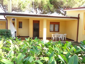 Un bâtiment jaune avec un patio couvert, une porte et une fenêtre avec des rideaux. On y trouve des chaises et une table en plastique, toutes deux blanches, sur le patio.