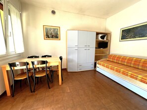 Et lille, godt oplyst rum med et træbord og stole, et hvidt klædeskab, en sofa med ternet betræk og to billeder i rammer på væggene.