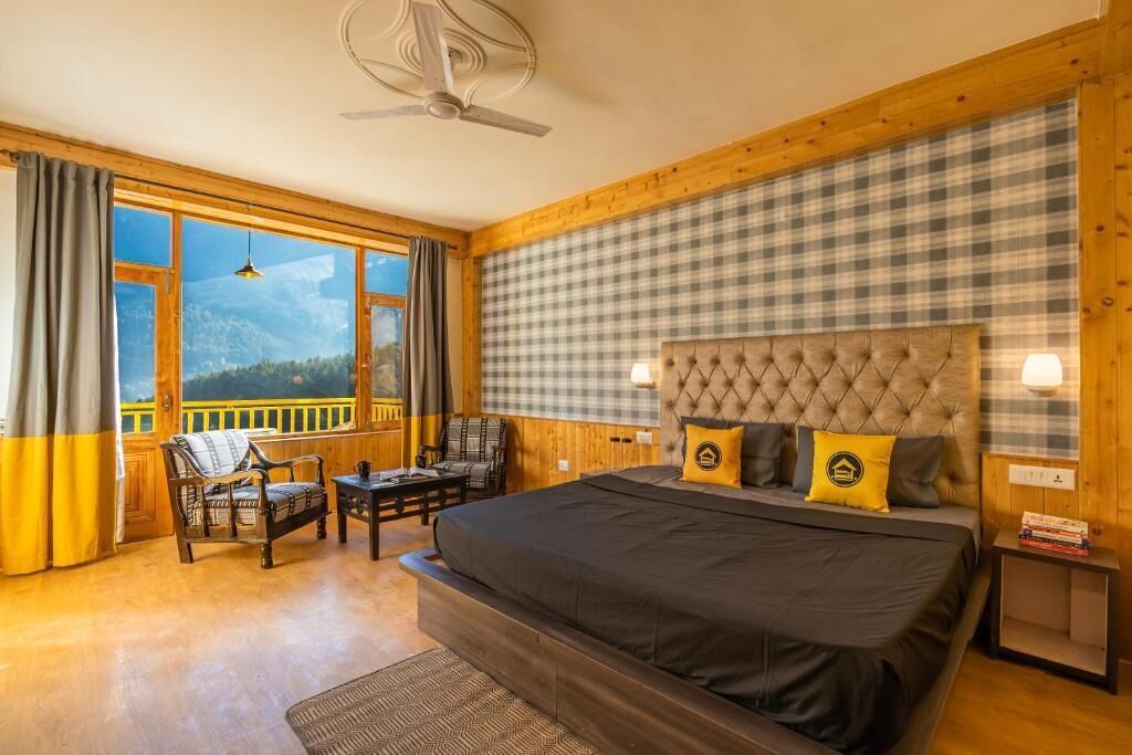 A bedroom with a large bed, a headboard, a window with curtains, a chair, a table, and a view of the mountains.