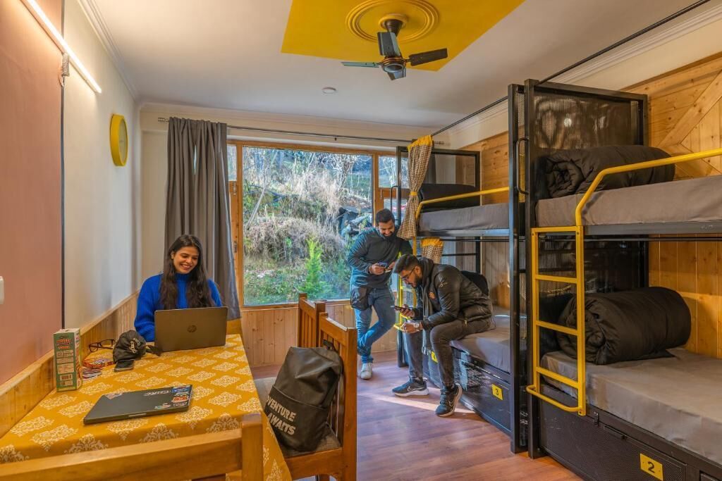 Een slaapzaal met stapelbedden, een tafel met een laptop en een raam met uitzicht op bomen.