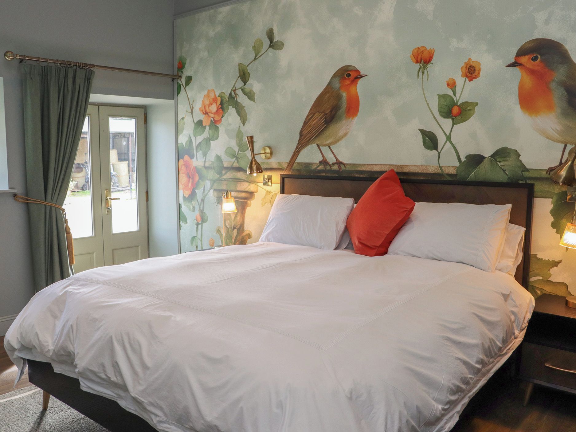 A bed with white linens, a red pillow, and a wall mural of birds and flowers.