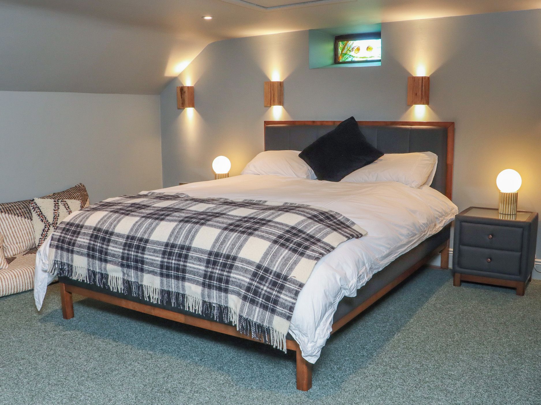 A bedroom with a bed, two bedside tables, a nightstand, a wall-mounted light, and a small window.